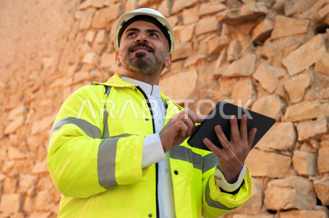 التعرف على فن العمارة القديمة، ذكرى تأسيس الدولة السعودية الأولى 22 فبراير، صورة مقربة لمهندس معماري عربي خليجي سعودي مبتسم يرتدي السترة والخوذة يستخدم جهاز التابلت في قصر سلوى في البجيري الدرعية، اساسيات البناء للبيوت والقصور التراثية، يوم بدينا 1727 م 