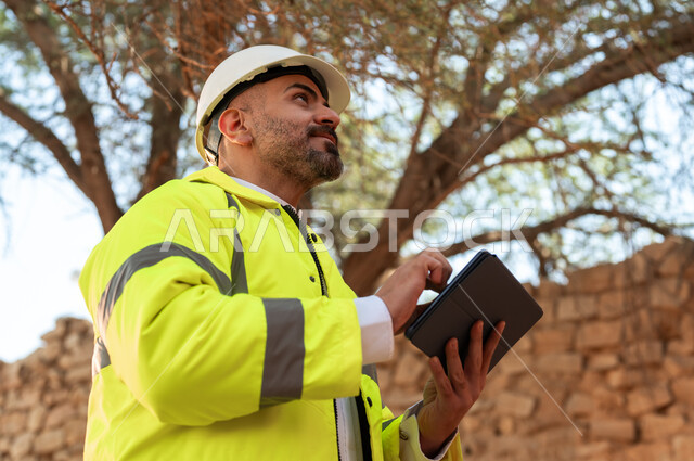 ذكرى تأسيس الدولة السعودية الأولى 22 فبراير، صورة مقربة لمهندس عربي خليجي سعودي مبتسم يرتدي السترة والخوذة يستخدم جهاز التابلت في قصر سلوى في البجيري الدرعية، اساسيات البناء للبيوت والقصور التراثية، يوم بدينا 1727 م، التعرف على فن العمارة القديمة 
