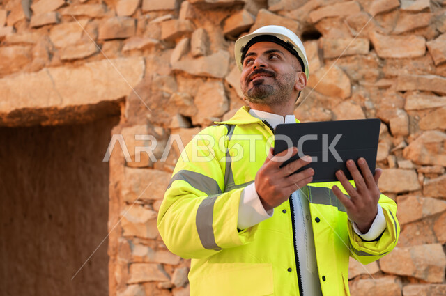 اساسيات البناء للبيوت والقصور التراثية، يوم بدينا 1727 م، التعرف على فن العمارة القديمة، ذكرى تأسيس الدولة السعودية الأولى 22 فبراير، صورة مقربة لمهندس معماري عربي خليجي سعودي مبتسم يرتدي السترة والخوذة يستخدم جهاز التابلت في قصر سلوى في البجيري الدرعية 