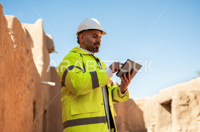 اطلال قصر سلوى في البجيري الدرعية، التعرف على فن العمارة القديمة واساسيات البناء للبيوت والقصور التراثية، يوم بدينا 1727 م، ذكرى تأسيس الدولة السعودية الأولى 22 فبراير، صورة مقربة لمهندس معماري عربي خليجي سعودي يرتدي السترة وخوذة الحماية يستخدم جهاز التابلت