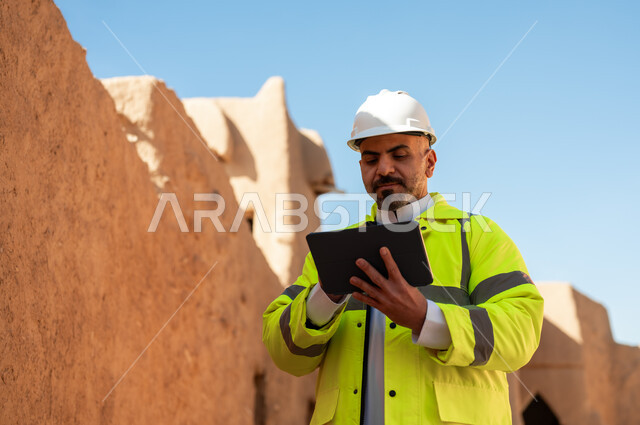 Getting to know the art of ancient architecture and the basics of construction for heritage houses and palaces, Day of Our Day 1727 AD, the anniversary of the founding of the first Saudi state, February 22, a close-up photo of a Saudi Gulf Arab architect wearing a jacket and a protective helmet using a tablet, the ruins of Salwa Palace in Al-Bujairi, Diriyah