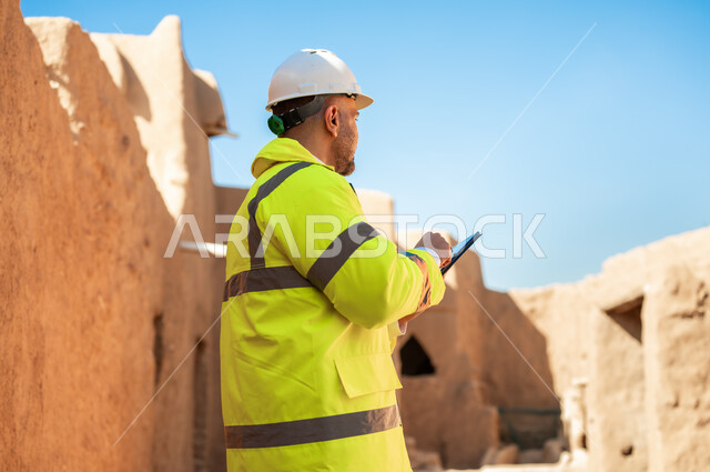 ذكرى تأسيس الدولة السعودية الأولى 22 فبراير، صورة مقربة لمهندس معماري عربي خليجي سعودي يرتدي السترة وخوذة الحماية يستخدم جهاز التابلت، اطلال قصر سلوى في البجيري الدرعية، التعرف على فن العمارة القديمة واساسيات البناء للبيوت والقصور التراثية، يوم بدينا 1727 م