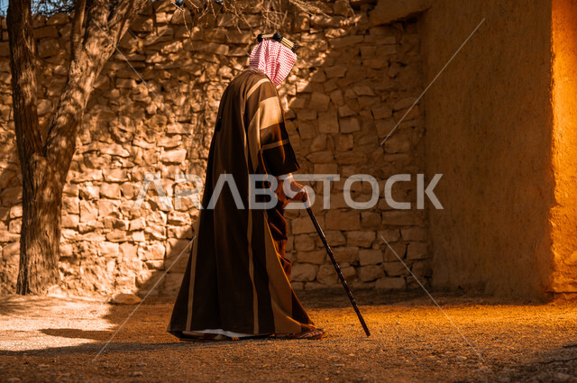 يوم بدينا 1727م، المباني الطينية الاثرية القديمة في الدرعية، تصوير مقرب لرجل عربي خليجي سعودي يرتدي الزي التراثي للمناسبات الوطنية والاحتفالية يستخدم العكاز في المشي، الجدران الطينية القديمة، باب خشبي اثري تاريخي، ذكرى تأسيس الدولة السعودية الاولى 22 فبراير