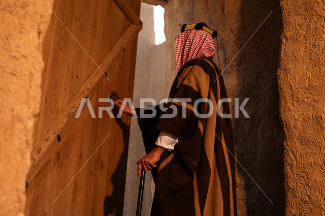 يوم بدينا 1727م، المباني الطينية الاثرية القديمة في الدرعية، تصوير مقرب لرجل عربي خليجي سعودي يرتدي الزي التراثي للمناسبات الوطنية والاحتفالية يقوم بفتح باب خشبي اثري تاريخي، الجدران الطينية القديمة، ذكرى تأسيس الدولة السعودية الاولى 22 فبراير