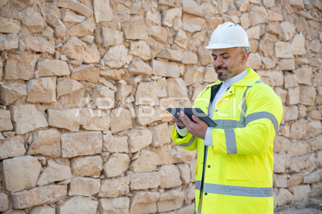Integrating technology and technology with the practical environment, monitoring and auditing work using a tape, Saudi professions and jobs, a Saudi Gulf Arab engineer wearing a helmet and work protection jacket standing in front of old buildings in Diriyah