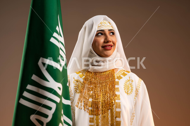 ذكرى يوم بدينا 1727 م، ارتداء زي المنطقة الشرقية في المناسبات الوطنية، بورتريه مقرب لإمرأة عربية خليجية سعودية مبتسمة ترتدي الشيلة والنشل تمسك علم تأسيس الدولة السعودية الأولى 22 فبراير تنظر الى شيء ما، التزين بالحلي والمجوهرات الذهبية التقليدية، خلفية بيج