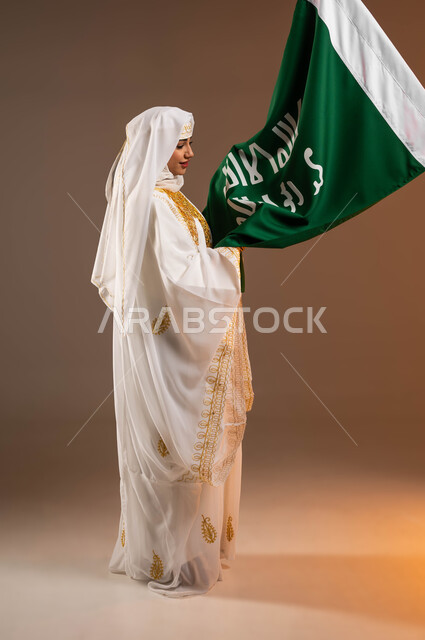 النظر الى علم تأسيس الدولة السعودية الأولى 22 فبراير، ذكرى يوم بدينا 1727 م، التزين بالحلي والمجوهرات الذهبية التقليدية، ارتداء زي المنطقة الشرقية في المناسبات الوطنية، بورتريه لإمرأة عربية خليجية سعودية مبتسمة ترتدي الشيلة والنشل، صورة الجسم بالكامل، خلفية بيج