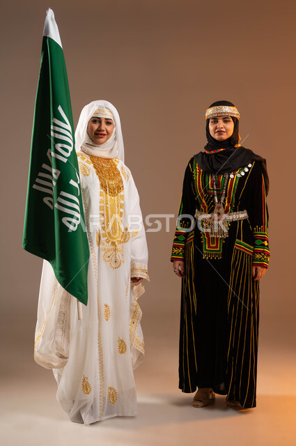 يوم بدينا 1727 م، بورتريه لإمرأتين عربيتين خليجيتين سعوديتين مبتسمتين ترتديان أزياء المنطقة الوسطى والشرقية تنظران الى الكاميرا بتعابير الفخر، الوقوف بجانب علم تأسيس الدولة السعودية الأولى في 22 فبراير، الاحتفال بالمناسبات الوطنية، صورة لكامل الجسم، خلفية بيج