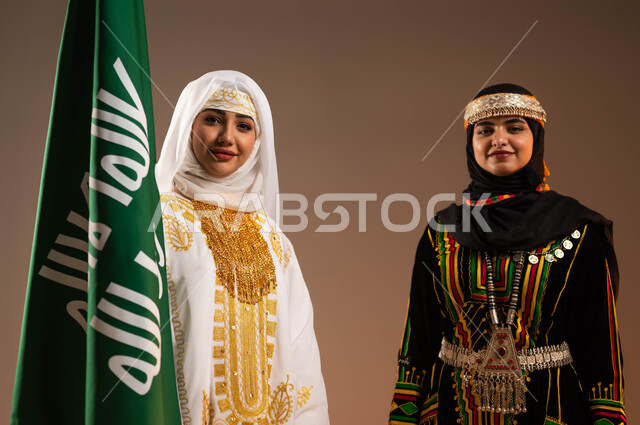 النظر الى الكاميرا بتعابير الفخر، ارتداء أزياء المنطقة الوسطى والشرقية في المناسبات الوطنية، يوم بدينا 1727 م، بورتريه مقرب لإمرأتين عربيتين خليجيتين سعوديتين مبتسمتين ترتديان الزي الشعبي تقفان بجانب علم تأسيس الدولة السعودية الأولى في 22 فبراير، خلفية بيج