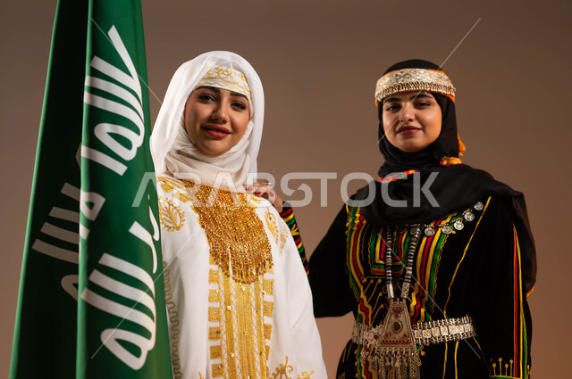 ارتداء أزياء المنطقة الوسطى والشرقية في المناسبات الوطنية، يوم بدينا 1727 م، بورتريه مقرب لإمرأتين عربيتين خليجيتين سعوديتين مبتسمتين ترتديان الزي الشعبي تقفان بجانب علم تأسيس الدولة السعودية الأولى في 22 فبراير، النظر الى الكاميرا بتعابير الفخر، خلفية بيج