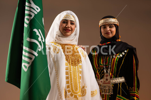 الوقوف بجانب علم تأسيس الدولة السعودية الأولى في 22 فبراير، ارتداء أزياء المنطقة الوسطى والشرقية في المناسبات الوطنية، يوم بدينا 1727 م، بورتريه مقرب لإمرأتين عربيتين خليجيتين سعوديتين مبتسمتين ترتديان الزي الشعبي تنظران الى الكاميرا بتعابير الفخر، خلفية بيج