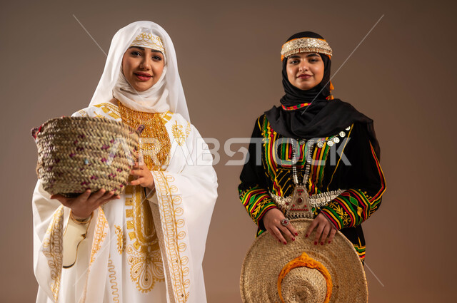 حرف ومهن شعبية نسائية، ذكرى تأسيس الدولة السعودية الأولى 22 فبراير، يوم بدينا 1727 م، بورتريه مقرب لإمرأتين عربيتين خليجيتين سعوديتين مبتسمتين ترتديان أزياء المنطقة الشرقية والوسطى تحملان مشغولات يدوية من القش، الاحتفال بالمناسبات الوطنية، خلفية بيج