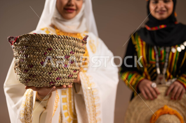 مهن وحرف شعبية نسائية، ذكرى تأسيس الدولة السعودية الأولى 22 فبراير، يوم بدينا 1727 م، بورتريه مقرب لإمرأتين عربيتين خليجيتين سعوديتين مبتسمتين ترتديان أزياء المنطقة الشرقية والوسطى تحملان مشغولات يدوية من القش، الاحتفال بالمناسبات الوطنية، خلفية بيج