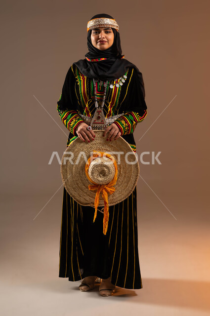 النظر باتجاه الكاميرا, مشغولات يدوية من القش، حرف ومهن شعبية نسائية، ذكرى تأسيس الدولة السعودية الأولى 22 فبراير، يوم بدينا 1727 م، بورتريه لإمرأة عربية خليجية سعودية ترتدي ازياء المنطقة الوسطى تحمل بيدها قبعة مصنوعة من القش، صورة لكامل الجسم، خلفية بيج