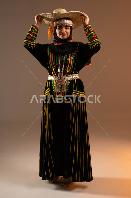 حرف ومهن شعبية نسائية, النظر باتجاه الكاميرا, مشغولات يدوية من القش، ذكرى تأسيس الدولة السعودية الأولى 22 فبراير، يوم بدينا 1727 م، بورتريه لإمرأة عربية خليجية سعودية ترتدي ازياء المنطقة الوسطى تضع على رأسها  قبعة مصنوعة من القش، صورة لكامل الجسم، خلفية بيج