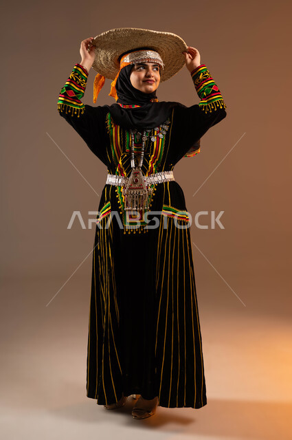 يوم بدينا 1727 م, مشغولات يدوية من القش، حرف ومهن شعبية نسائية، ذكرى تأسيس الدولة السعودية الأولى 22 فبراير، بورتريه لإمرأة عربية خليجية سعودية ترتدي ازياء المنطقة الوسطى تضع على رأسها قبعة مصنوعة من القش, النظر الى شيء ما، صورة لكامل الجسم، خلفية بيج