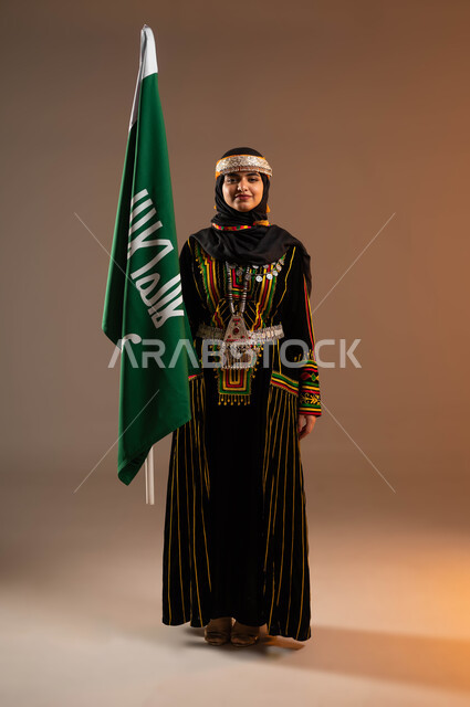 يوم بدينا ١٧٢٧م, الوقوف باستقامة والنظر الى الكاميرا, الاعتزاز بانجازات البلاد، حمل علم التأسيس بشموخ، الاحتفال بذكرى تأسيس الدولة السعودية الاولى ٢٢فبراير، بورتريه مقرب لشابة عربية خليجية سعودية ترتدي زي المنطقة الوسطى, صورة الجسم بالطول الكامل، خلفية بيج