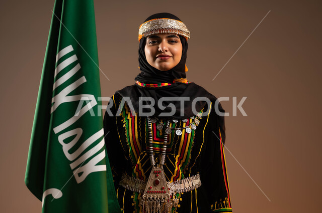 الوقوف باستقامة والنظر الى الكاميرا, الاعتزاز بانجازات البلاد، ارتداء الازياء الشعبية، حمل علم التأسيس بشموخ، الاحتفال بذكرى تأسيس الدولة السعودية الاولى ٢٢فبراير، بورتريه مقرب لشابة عربية خليجية سعودية ترتدي زي المنطقة الوسطى، يوم بدينا ١٧٢٧م، خلفية بيج