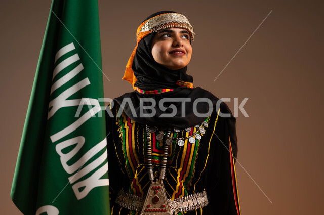 الاعتزاز بانجازات البلاد, النظر الى شيء ما، ارتداء الازياء الشعبية في المناسبات الوطنية، حمل علم التأسيس بشموخ، الاحتفال بذكرى تأسيس الدولة السعودية الاولى ٢٢فبراير، بورتريه مقرب لشابة عربية خليجية سعودية ترتدي زي المنطقة الوسطى، يوم بدينا ١٧٢٧م، خلفية بيج