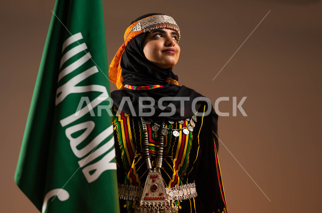 النظر الى شيء ما, الاعتزاز بانجازات البلاد، ارتداء الازياء الشعبية في المناسبات الوطنية، حمل علم التأسيس بشموخ، الاحتفال بذكرى تأسيس الدولة السعودية الاولى ٢٢فبراير، بورتريه مقرب لشابة عربية خليجية سعودية ترتدي زي المنطقة الوسطى، يوم بدينا ١٧٢٧م، خلفية بيج