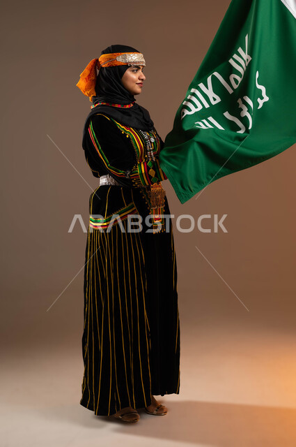 النظر الى علم تأسيس الدولة السعودية الأولى 22 فبراير، ذكرى يوم بدينا 1727 م، ارتداء زي المنطقة الوسطى في المناسبات الوطنية، بورتريه لإمرأة عربية خليجية سعودية مبتسمة تمسك بيدها علم التأسيس, الاعتزاز بالبلد والانتماء اليه، صورة الجسم بالطول الكامل، خلفية بيج