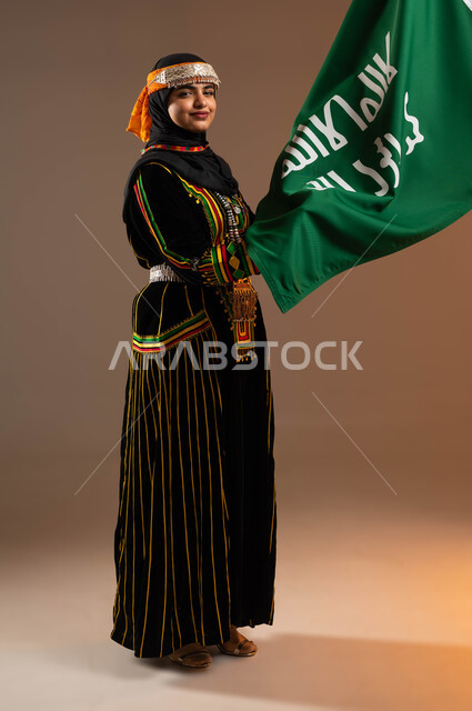 النظر باتجاه الكاميرا, ذكرى تأسيس الدولة السعودية الأولى 22 فبراير، يوم بدينا 1727 م، بورتريه لإمرأة عربية خليجية سعودية مبتسمة ترتدي زي المنطقة الوسطى تمسك بيدها علم التأسيس, الاعتزاز بالبلد والانتماء اليه، صورة الجسم بالطول الكامل، خلفية بيج