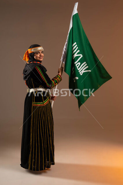 رفع علم التأسيس بإيماءات الثقة و الفخر بانجازات الوطن، الأزياء الشعبية التقليدية، بورتريه لامرأة عربية خليجية سعودية ترتدي زي المنطقة الوسطى، ذكرى تأسيس الدولة السعودية الأولى ٢٢ فبراير، مفهوم حب الوطن والارتباط به، يوم بدينا١٧٢٧م، صورة لكامل الجسم, خلفية بيج