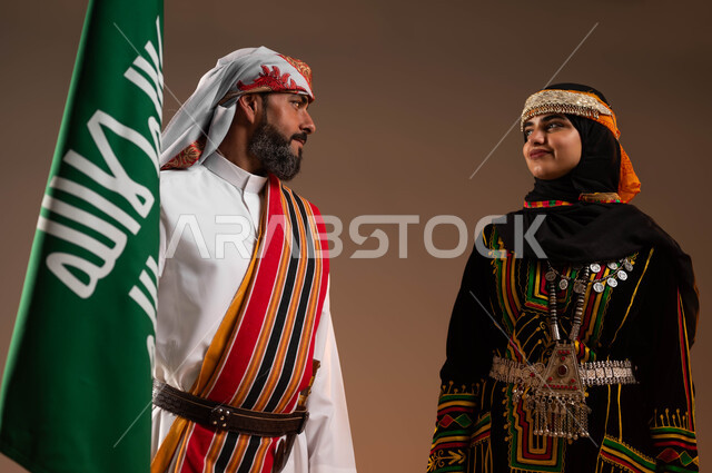 النظر الى بعض بتباهي, الوقوف بجانب علم التأسيس بثقة وفخر, توارث حب الوطن من الأجداد, بورتريه مقرب لرجل وامرأة عربيان خليجيان سعوديان يرتديان ازياء المنطقة الجنوبية والوسطى, يوم بدينا 1727م, ذكرى تأسيس الدولة السعودية الأولى في 22 فبراير, خلفية بيج