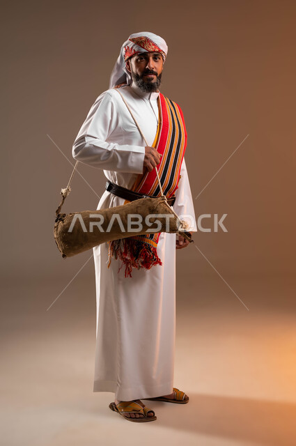 ذكرى تأسيس الدولة السعودية الأولى 22فبراير, ارتداء أزياء المنطقة الجنوبية, يوم بدينا 1727 م، بورتريه لرجل عربي خليجي سعودي يرتدي الثوب والغترة والشال الجنوبي ويحمل قربة الماء للسقي، استخدام الادوات الشعبية القديمة، صورة الجسم بالطول الكامل، خلفية بيج