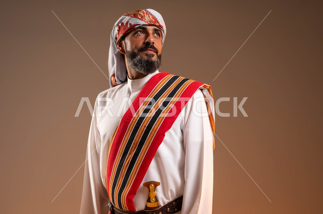 الوقوف باستقامة والنظر الى شيء ما, يوم بدينا 1727م، ارتداء أزياء المنطقة الجنوبية للاحتفال في المناسبات الوطنية، بورتريه مقرب لرجل عربي خليجي سعودي مبتسم يرتدي الثوب والغترة مع الشال الجنوبي، ذكرى تأسيس الدولة السعودية الأولى 22فبراير، خلفية بيج