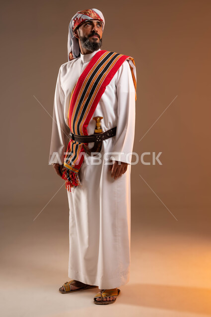 يوم بدينا 1727م، الوقوف باستقامة بإيماءات الفخر والاعتزاز, ارتداء أزياء المنطقة الجنوبية للاحتفال في المناسبات الوطنية، بورتريه لرجل عربي خليجي سعودي مبتسم يرتدي الثوب والغترة مع الشال الجنوبي، ذكرى تأسيس الدولة السعودية الأولى 22فبراير، صورة الجسم بالكامل، خلفية بيج