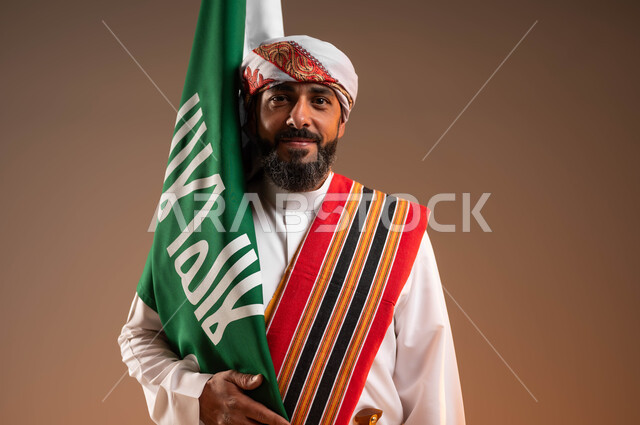 الوقوف باستقامة والنظر إلى الكاميرا بإيماءات الفرح في يوم بدينا 1727م، مشاعر الحماس أثناء الاحتفال بذكرى تأسيس الدولة العربية السعودية 22فبراير، بورتريه مقرب لرجل عربي خليجي سعودي يرتدي الثوب والغترة مع الشال الجنوبي يحمل بيده علم التأسيس، خلفية بيج