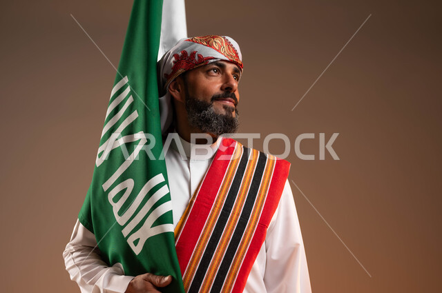 الاحتفال باليوم الوطني السعودي 22فبراير, النظر الى شيء ما بفخر، يوم بدينا 1727م، بورتريه مقرب لرجل عربي خليجي سعودي يرتدي الثوب والغترة مع الشال الجنوبي يحمل بيده علم يوم التأسيس, ارتداء ازياء المنطقة الجنوبية, خلفية بيج