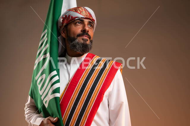 النظر الى شيء ما بفخر, الاحتفال باليوم الوطني السعودي 22فبراير، يوم بدينا 1727م، بورتريه مقرب لرجل عربي خليجي سعودي يرتدي الثوب والغترة مع الشال الجنوبي يحمل بيده علم يوم التأسيس, ارتداء ازياء المنطقة الجنوبية, خلفية بيج