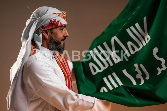 المناسبات والاحتفالات الوطنية, ارتداء ازياء المنطقة الجنوبية، بورتريه مقرب من الجانب لمواطن عربي خليجي سعودي يرتدي الثوب والغترة مع الشال الجنوبي ويحمل بيده علم التأسيس بسرور وسعادة، يوم العلم السعودي 22فبراير، يوم بدينا 1727م، خلفية بيج
