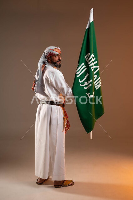 مشاعر الحماس أثناء الاحتفال باليوم الوطني السعودي 22فبراير، يوم بدينا 1727م، بورتريه من الخلف لرجل عربي خليجي سعودي يرتدي الثوب والغترة مع الشال الجنوبي يحمل بيده علم يوم التأسيس, ارتداء ازياء المنطقة الجنوبية, صورة الجسم بالطول الكامل, خلفية بيج