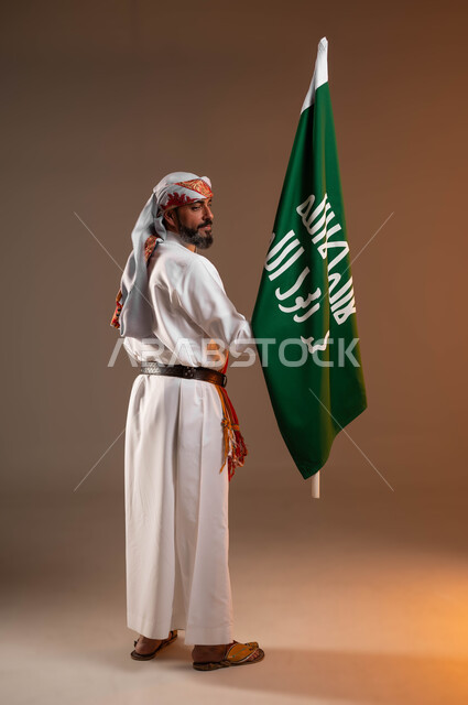 المشاركة بالمناسبات الوطنية, يوم بدينا 1727م، مشاعر الحماس أثناء الاحتفال باليوم الوطني السعودي 22فبراير، بورتريه من الخلف لرجل عربي خليجي سعودي يرتدي الثوب والغترة يحمل بيده علم يوم التأسيس, ارتداء ازياء المنطقة الجنوبية, صورة الجسم بالطول الكامل, خلفية بيج