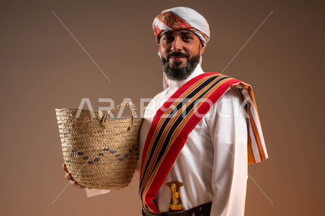 ارتداء ازياء المنطقة الجنوبية, النظر الى الكاميرا بإيماءات الفخر , حرف ومهن يدوية قديمة، ذكرى تأسيس الدولة السعودية 22فبراير، يوم بدينا 1727م، بورتريه مقرب مبتسم لرجل عربي خليجي سعودي يرتدي الثوب والغترة مع الشال الجنوبي يمسك بيده سلة مصنوعة من القش، خلفية بيج