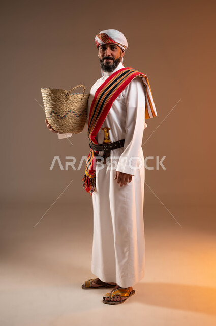 النظر الى الكاميرا بإيماءات التباهي والفخر , حرف ومهن يدوية قديمة، ذكرى تأسيس الدولة السعودية 22فبراير، يوم بدينا 1727م، بورتريه لرجل عربي خليجي سعودي يرتدي الثوب والغترة مع الشال الجنوبي يمسك بيده سلة مصنوعة من القش, صورة الجسم بالطول الكامل، خلفية بيج