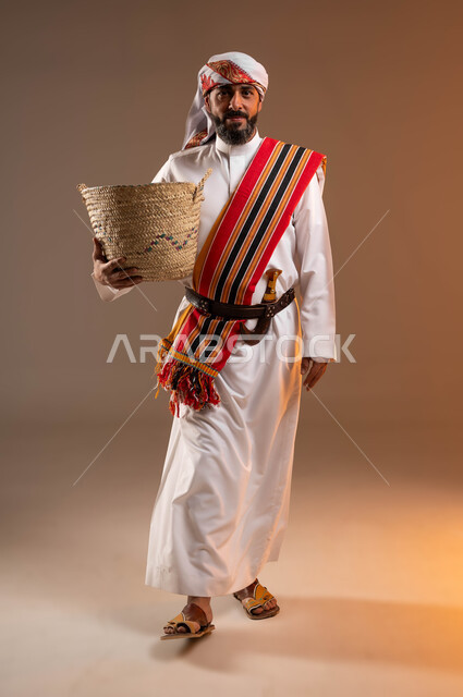 حرف ومهن يدوية قديمة, النظر الى الكاميرا بإيماءات التباهي، ذكرى تأسيس الدولة السعودية 22فبراير، يوم بدينا 1727م، بورتريه لرجل عربي خليجي سعودي يرتدي الثوب والغترة مع الشال الجنوبي يمسك بيده سلة مصنوعة من القش, صورة الجسم بالطول الكامل، خلفية بيج
