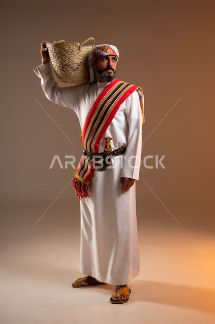 حرف ومهن يدوية قديمة, النظر الى شيء ما، ذكرى تأسيس الدولة السعودية 22فبراير، يوم بدينا 1727م، ارتداء أزياء المنطقة الجنوبية، بورتريه لرجل عربي خليجي سعودي يرتدي الثوب والغترة مع الشال الجنوبي يضع على كتفه سلة مصنوعة من القش, صورة الجسم بالطول الكامل، خلفية بيج
