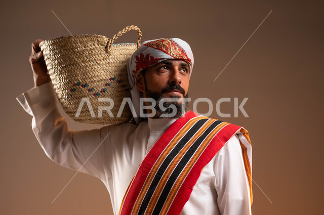 النظر الى شيء ما, حرف ومهن يدوية قديمة، ذكرى تأسيس الدولة السعودية 22 فبراير، يوم بدينا 1727 م، ارتداء أزياء المنطقة الجنوبية، بورتريه مقرب لرجل عربي خليجي سعودي يرتدي الثوب والغترة مع الشال الجنوبي يضع على كتفه سلة مصنوعة من القش، خلفية بيج