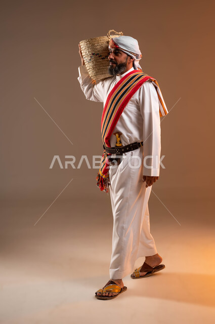 ذكرى تأسيس الدولة السعودية 22 فبراير، حرف ومهن يدوية قديمة، يوم بدينا 1727 م، ارتداء أزياء المنطقة الجنوبية، بورتريه من الجانب لرجل عربي خليجي سعودي يرتدي الثوب والغترة مع الشال الجنوبي يمشي ويضع على كتفه سلة مصنوعة من القش، صورة الجسم بالكامل، خلفية بيج