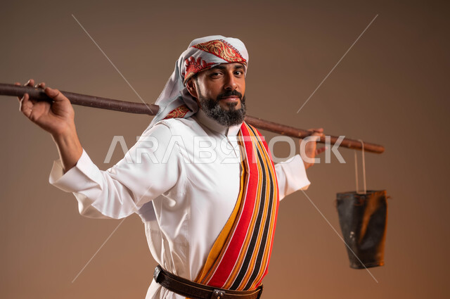 ارتداء ازياء المنطقة الجنوبية، تأسيس الدولة السعودية الاولى 22فبراير، سقاي الماء يقوم بالتجول لسقي الناس من البئر، يوم بدينا 1727م، بورتريه مقرب لرجل عربي خليجي سعودي يرتدي الثوب والغترة والشال الجنوبي يحمل دلو من الجلد, خلفية بيج