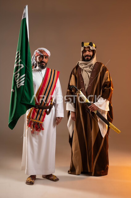 ذكرى تأسيس الدولة السعودية الأولى في 22 فبراير يوم بدينا 1727م, بورتريه لرجلان عربيات خليجيان سعوديان مبتسمان يرتديان أزياء المنطقة الوسطى والجنوبية, يمسكان بيدهما علم التأسيس والسيف وينظران إلى الكاميرا, صورة الجسم بالطول الكامل, خلفية بيج