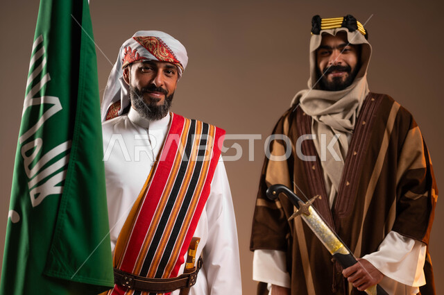 Pride and pride in the country, the day of our birth 1727 AD, a close-up portrait of two smiling Saudi Gulf Arab men wearing the fashions of the central and southern regions, holding the founding flag and sword in their hands and looking at the camera, the anniversary of the founding of the first Saudi state on February 22, beige background