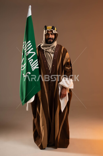 Looking at the camera with gestures of pride and pride in the Arab identity, a portrait of a Saudi Gulf Arab man wearing a golden iqal, dhikla, and ghutra, raising the founding flag with ostentation and pride, the anniversary of the founding of the first Saudi state in 1727 AD, Dayna Day, February 22, full body portrait, beige background