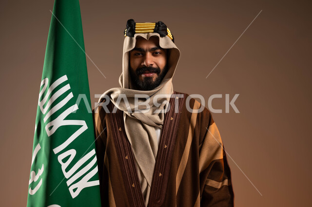 ايماءات الفخر والاعتزاز بالهوية العربية، بورتريه مقرب لرجل عربي خليجي سعودي مبتسم يرتدي العقال الذهبي والدقلة والغترة، رفع علم التأسيس بتباهي واعتزاز، المشاركة بالاحتفالات والمناسبات الوطنية، ذكرى تأسيس الدولة السعودية الأولى١٧٢٧م،يوم بدينا٢٢فبراير، خلفية بيج