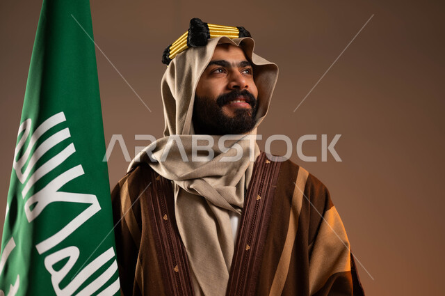 الفخر والاعتزاز بالهوية العربية، يوم بدينا ٢٢فبراير، المشاركة بالاحتفالات الوطنية، بورتريه مقرب لرجل عربي خليجي سعودي مبتسم يرتدي العقال الذهبي والدقلة والغترة ينظر لمكان ما بانتباه، رفع علم التأسيس بتباهي واعتزاز، ذكرى تأسيس الدولة السعودية الأولى١٧٢٧م، خلفية بيج