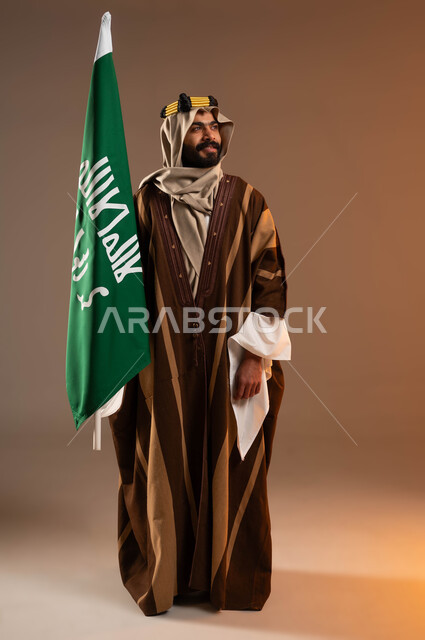 ذكرى تأسيس الدولة السعودية الأولى١٧٢٧م، المشاركة بالمناسبات و الاحتفالات الوطنية، رفع علم التأسيس بفخر وتباهي، يوم بدينا ٢٢فبراير،  بورتريه لرجل عربي خليجي سعودي مبتسم يرتدي العقال الذهبي والدقلة والغترة ينظر إلى الجانب بانتباه، صورة لكامل الجسم، خلفية بيج